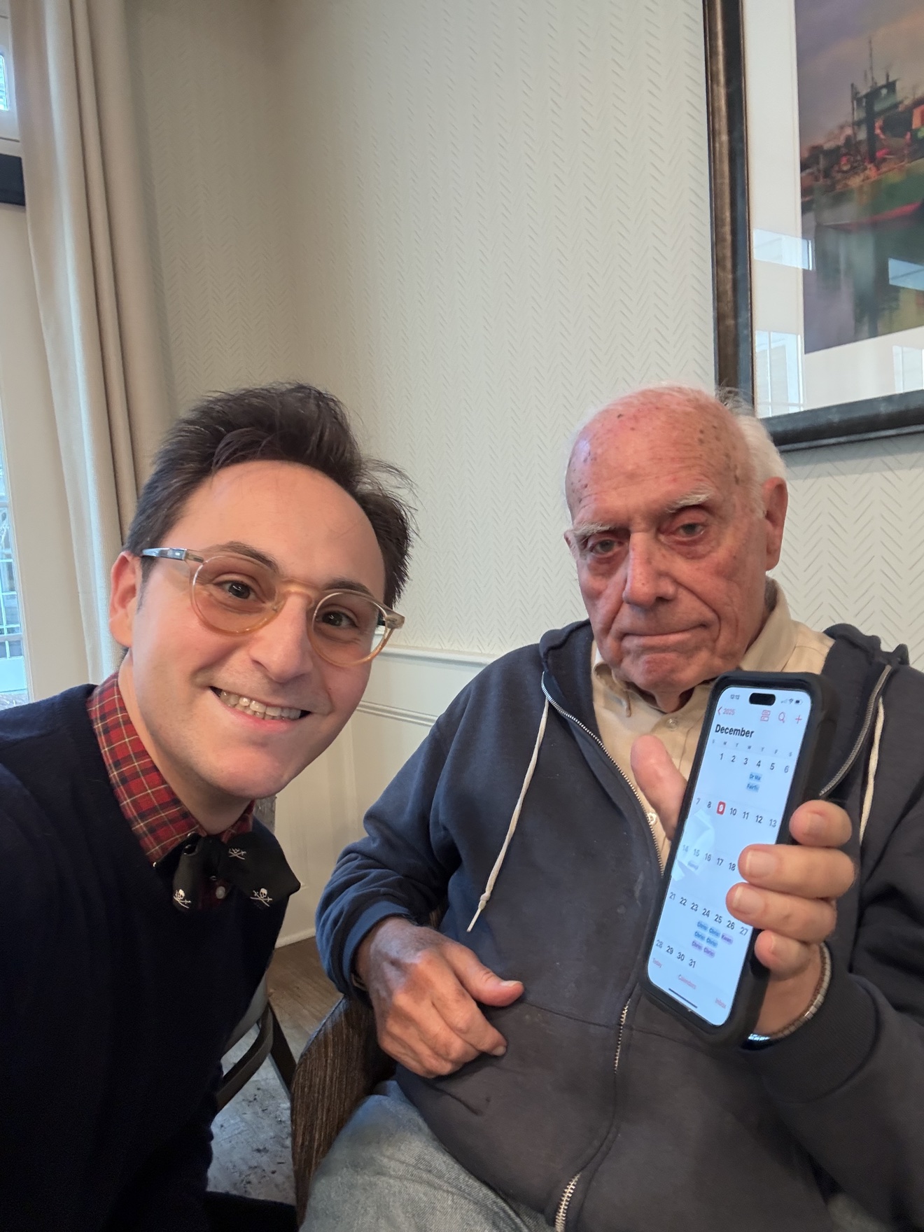 Selfie of two smiling people, one holding up an iPhone showing a calendar app, in a bright senior-living lounge